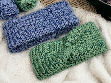Two speckled knit-look headbands, one blue and one green with a twisted center, resting on a cream blanket with yarn balls nearby.
