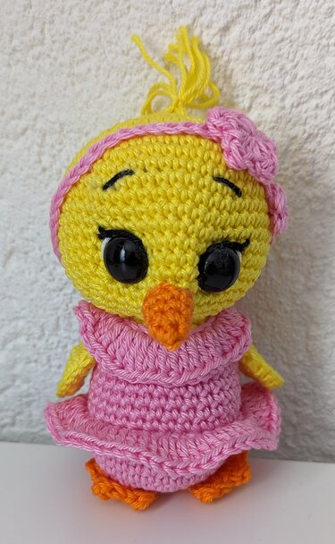 Gehäkeltes gelbes Amigurumi-Küken mit rosa Rüschenkleid, rosa Stirnband mit Blüte sowie orangefarbenem Schnabel und Füßen vor hellem Hintergrund.