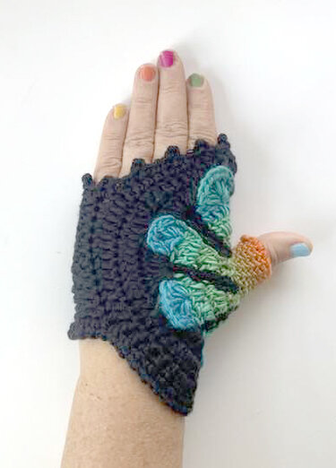 Hand mit dunkelgrauer gehäkelter fingerloser Stulpe und blau-grüner Schmetterlings-Applikation, bunte Fingernägel
