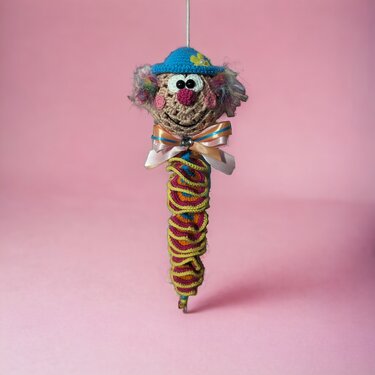 gehäkelter Clown‑Anhänger mit blauem Häubchen und buntem Spiralkörper vor rosa Hintergrund