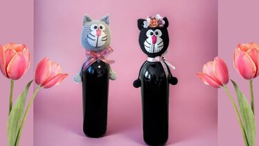 Zwei Flaschen mit gehäkelten Katzenköpfen vor rosa Hintergrund, links grau mit rosa Schleife, rechts schwarz mit Blumenkrone.