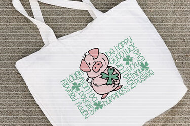 Weiße Stofftasche mit rosa Schwein, das ein Kleeblatt hält, umgeben von englischen Glückswörtern