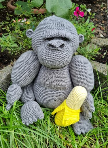 Gehäkelter grauer Amigurumi-Affe sitzt im Gras und hält eine gelbe gehäkelte Banane