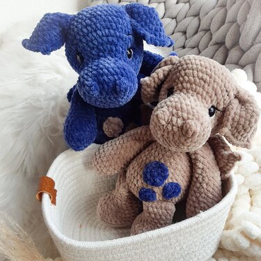Zwei gehäkelte Amigurumi-Dinos, ein blauer und ein beiger mit blauen Punkten, in einem weißen Korb.