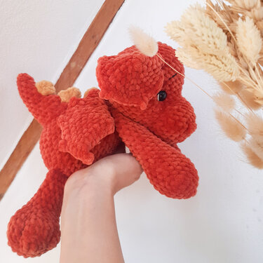 Hand hält einen rostroten, gehäkelten Amigurumi‑Drachen mit Flügeln, Rückenstacheln und Horn.