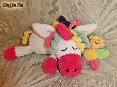 Gehäkeltes weißes Einhorn mit pinker Schnauze und pastellfarbener Mähne, liegend mit geschlossenen Augen neben kleinem Schlafengel