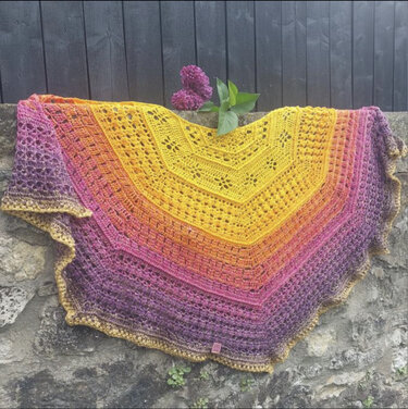 Gehäkeltes Dreieckstuch in Gelb‑Orange‑Violett mit Spitzen und gezacktem Rand, auf Steinmauer drapiert