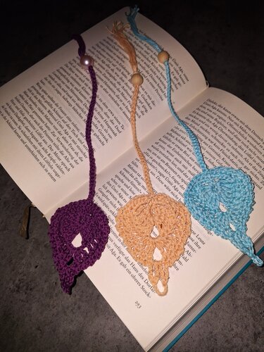 Drei handgehäkelte Blattform-Lesezeichen (Lila, Beige, Hellblau) mit Perlen in einem aufgeschlagenen Buch.