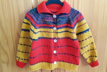 Gestrickte Kinderjacke mit bunten Streifen, rotem Kragen und weißen Knöpfen auf einem blauen Kleiderbügel