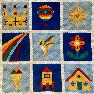 Gehäkelte Babydecke mit neun Quadraten und bunten Pixelmotiven wie Krone, Heißluftballon und Rakete.