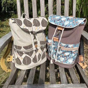 Zwei Stoffrucksäcke mit Klappen, Reißverschlusstaschen und Kordelzug auf einer Holzbank