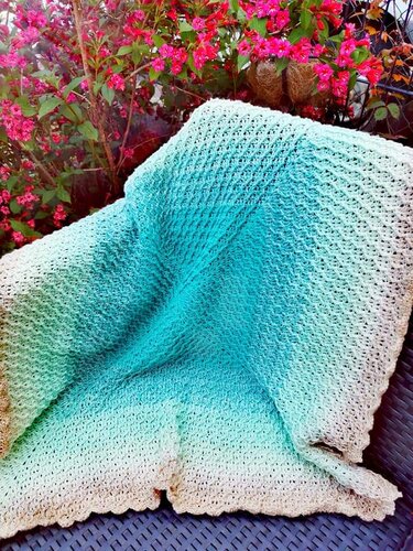 Gehäkelte Decke mit Ombre-Farbverlauf in Türkis, Mint und Beige vor pinken Blüten