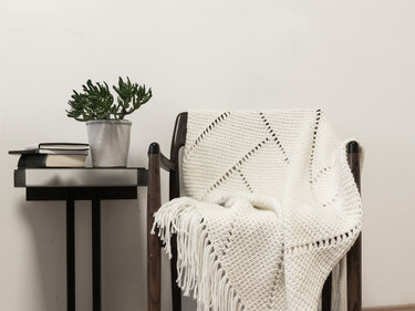 Cream crocheted blanket with geometric openwork pattern and fringe draped over a wooden chair next to a small table with a succulent and books.