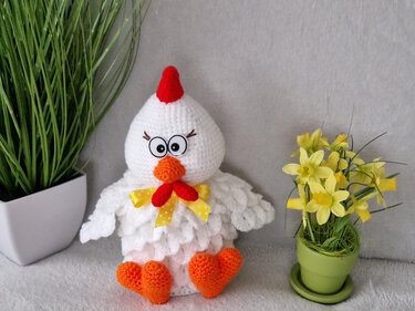 Weißes Amigurumi-Osterhuhn mit rotem Kamm, orangefarbenem Schnabel und herzförmigen Füßen neben Narzissen