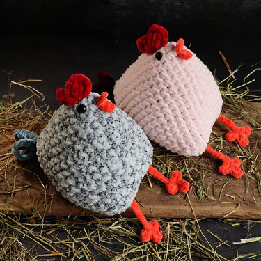 Pair of handmade amigurumi chickens in pink and grey with red combs, on wood and hay.