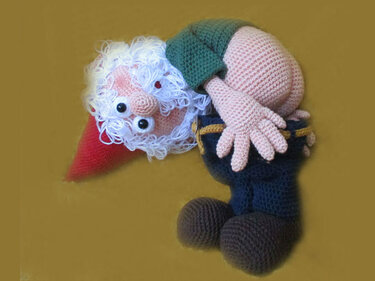 Crocheted amigurumi gnome with red hat, curly white beard, green top, blue pants, and brown shoes on a yellow background.