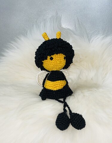 Gehäkelte schwarz-gelbe Amigurumi-Biene mit Lockenkranz und weißen Flügeln auf flauschigem Stoff.