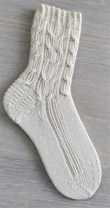 Cremefarbene handgestrickte Damensocke mit Zopfmuster am Schaft, gerippter Ferse und strukturierter Spannpartie auf hellem Holzuntergrund.