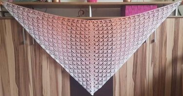 Gehäkeltes Dreieckstuch mit rosa-cremefarbenem Farbverlauf und Blattmuster, aufgehängt vor Holzschrank