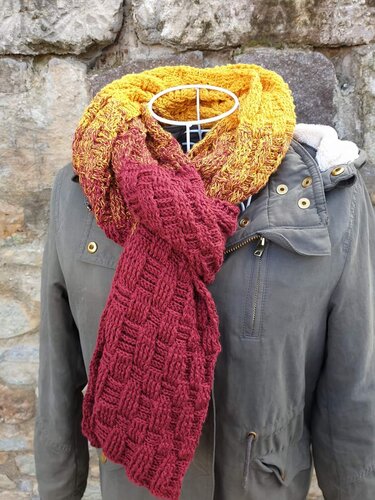 Gehäkelter Schal in Gelb, Orange und Rot mit strukturiertem Muster, drapiert an einer Schaufensterpuppe vor einer Steinwand.