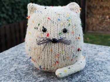 Gestrickte cremefarbene Katze mit bunten Sprenkeln, schwarzen Perlenaugen und grauen Schnurrhaaren auf grauem Untergrund