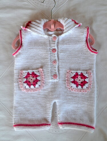 Weißer gestrickter Babystrampler mit rosa Granny‑Square‑Taschen und rosa Knöpfen an einem rosafarbenen Kleiderbügel