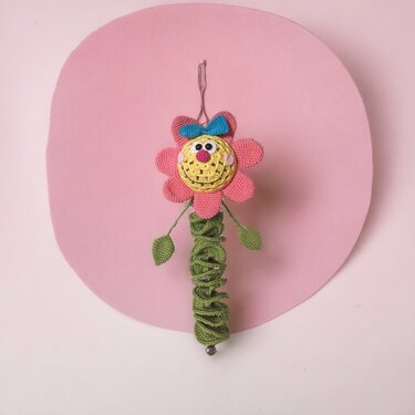 Gehäkelte hängende Blume mit rosa Blüten, gelbem Gesicht, blauem Schleifchen und grünem Spiralstiel