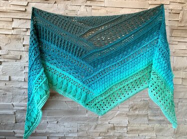 Ombre crocheted triangle shawl in a teal-to-aqua gradient, showing alternating lace and textured stitch sections, draped on a light stone wall.