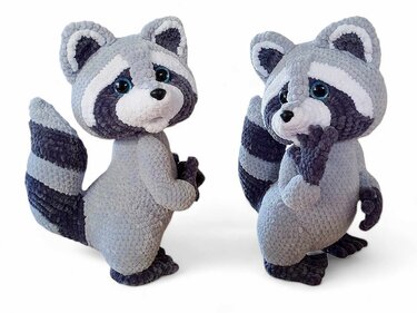 Two crocheted amigurumi raccoons with striped tails and facial masks, one facing forward and one with a paw near its face.