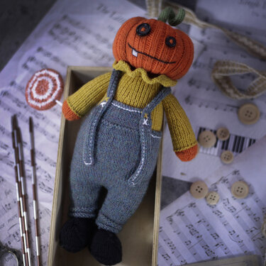 Knitted pumpkin amigurumi wearing mustard ruffle sweater and gray overalls holding a lollipop