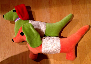 Zwei handgenähte Dackel-Kuscheltiere (grün-rot und orange-grün) auf Holzboden