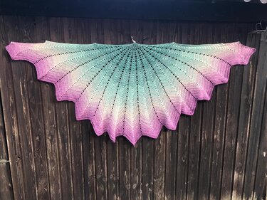 Fächerförmiges gehäkeltes Tuch mit Farbverlauf in Pink, Weiß und Türkis, aufgehängt vor dunkler Holzwand.