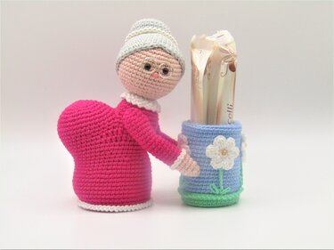 Gehäkelte pinke Oma-Amigurumi-Figur mit Brille und Herzbody hält blaues gehäkeltes Behältnis mit weißen Blumen