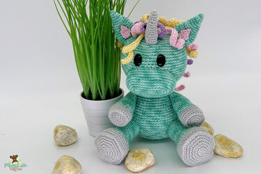 Mintgrünes gehäkeltes Einhorn-Amigurumi mit grauen Hufen, grauem Horn und bunter Lockenmähne neben einer Topfpflanze.