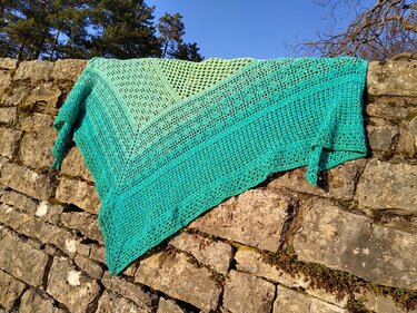 Gehäkeltes Dreieckstuch in Hellgrün bis Türkis mit Ajour- und Streifenmuster auf einer Steinmauer