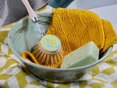 Mustard-yellow knitted potholder with raised square stitch pattern in a ceramic bowl with wooden dish brush and bar of soap