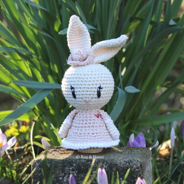Gehäkelter Amigurumi-Hase im pastellrosa Kleid mit Blütenhaarband vor grünen Blättern und Krokussen