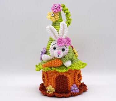 Gehäkelter Amigurumi-Hase mit rosa Schleife, der aus einem grünen und orangefarbenen Häuschen mit Blumen und Karotte hervorschaut.