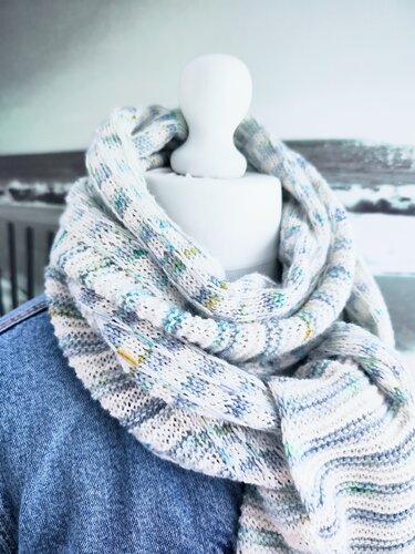 Heller gestrickter Loop-Schal mit blauen, grünen und gelben Sprenkeln, an einer Schaufensterpuppe über einer Jeansjacke drapiert.