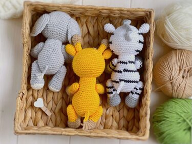 Gehäkelte Amigurumi-Tiere: grauer Elefant, gelbe Giraffe, gestreiftes Zebra in einem Flechtkorb, daneben Garnknäuel.