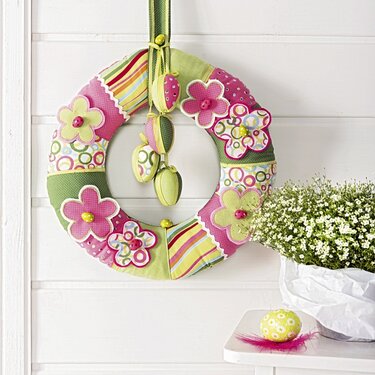Bunter Stoffkranz in Patchwork-Optik mit Applikationsblumen, hängenden Stoff-Ostereiern und grüner Aufhängeschleife an einer weißen Wand