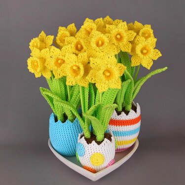 Yellow crocheted daffodil flowers with green leaves in egg-shaped crochet pots on a heart-shaped tray