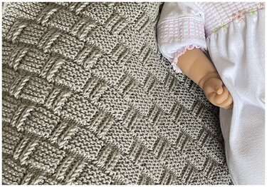 Nahaufnahme eines gestrickten K	 rbchenmusters auf einer beigen Babydecke; am Bildrand eine Puppe mit wei	 em Outfit