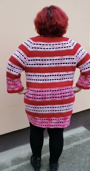Rückenansicht einer Person mit rotem Haar, die eine gestreifte Häkeljacke in Rot- und Rosatönen mit offenem Maschenbild trägt.