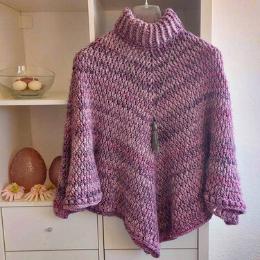 Gehäkelter Poncho in lila-melierten Tönen mit hohem geripptem Rollkragen an einem Kleiderbügel, mit Quastenanhänger.