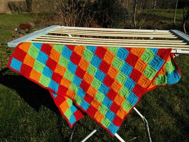 Gehäkelte Decke mit Rautenmuster in Rot, Orange, Hellgrün und Hellblau, aufgehängt auf einem Wäscheständer im Garten.