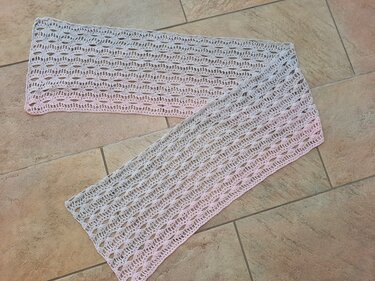 Gehäkelter Schal mit wiederholendem Loch- und Muschelmuster, helles Garn, auf Fliesen liegend