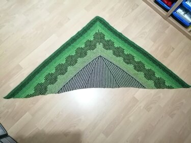 Gestricktes grünes Dreieckstuch mit keltischem Knotenmuster und Streifen, flach auf hellem Holzboden