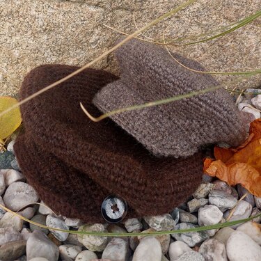 Herbstliches Stillleben: zwei gehäkelte Mützen in Braun und Taupe auf Kies mit getrockneten Blättern