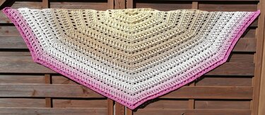 Gehäkeltes halbrundes Tuch mit Farbverlauf von Gelb zu Rosa, Lochmuster und pinker Kante, aufgehängt vor Holzlatten